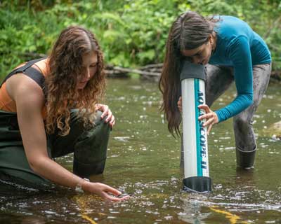 A female student in green pants partially kneels down and reaches her hand into a river and a second student in a blue shirt bends over and uses a tube to look beneath the surface of the river.