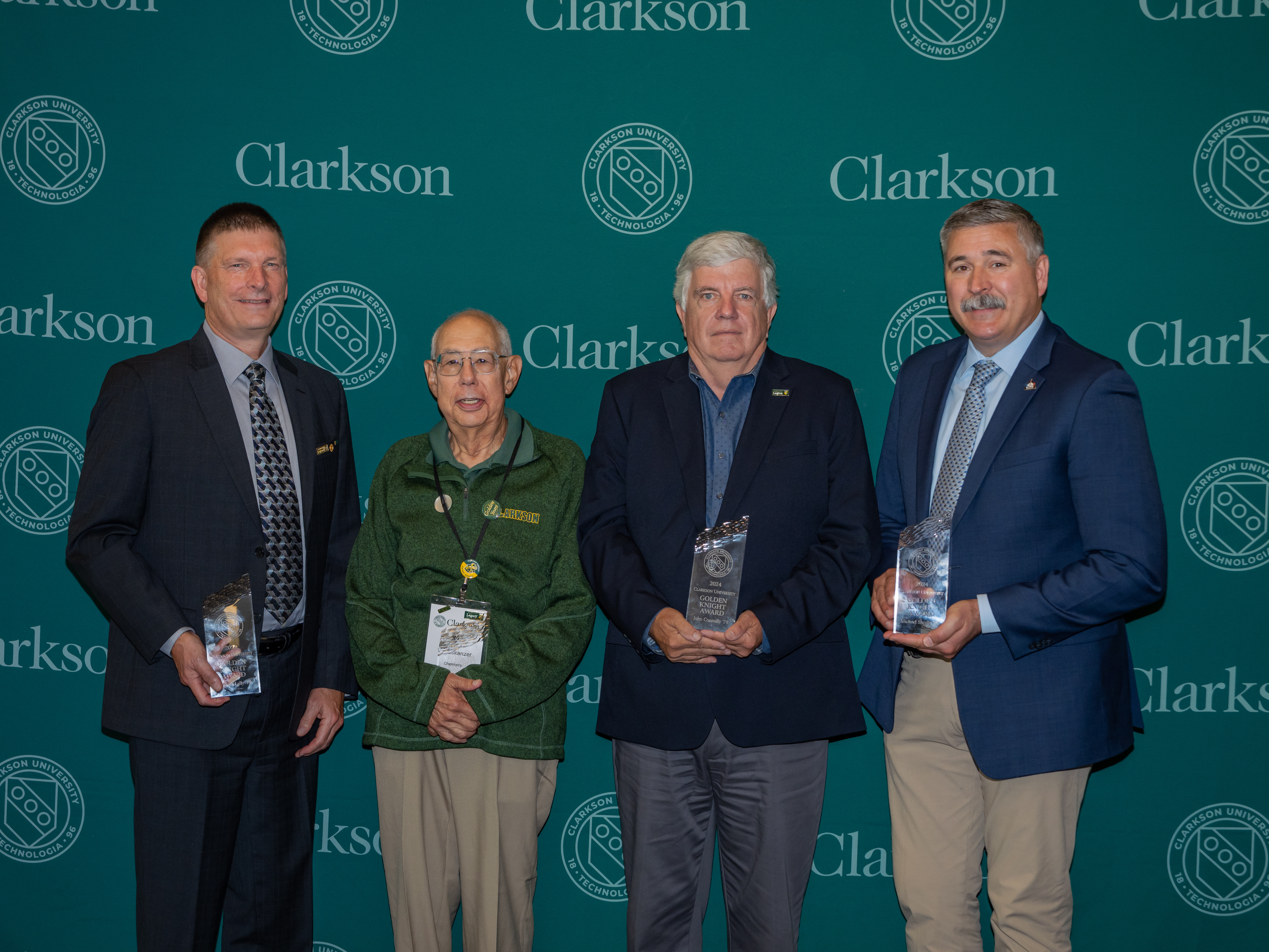 Four men pose for photos with crystal plaques in front of a media banner with Clarkson University logos