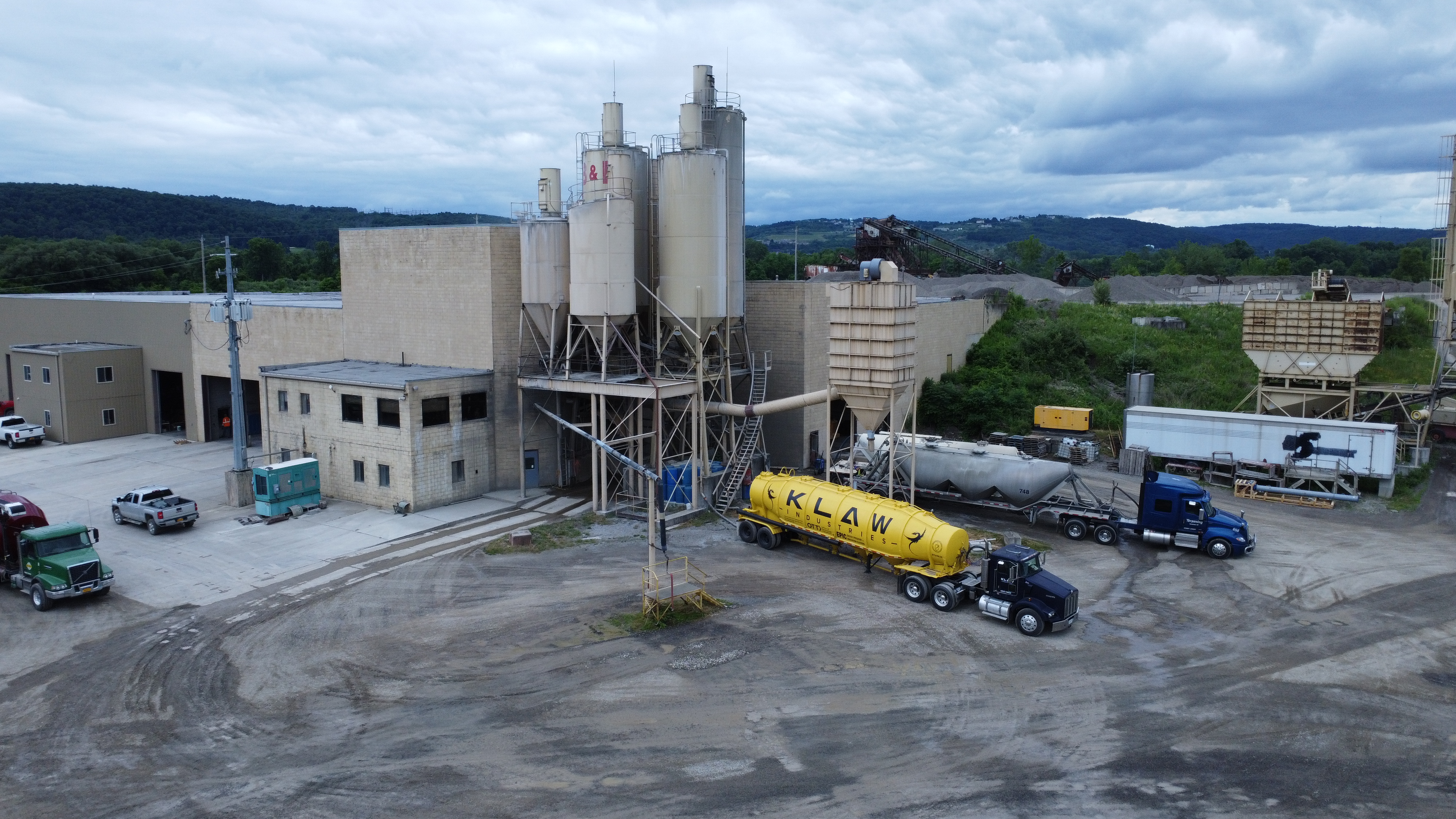 A yellow trailer on a semi-truck with KLAW Industries printed on it is backed up to an industrial facility in a large dirt parking lot. 