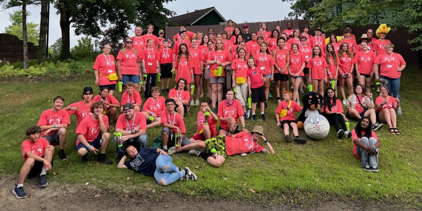 Dozens of students and camp counselors in pink shirts pose for a photo on a grassy hillside.