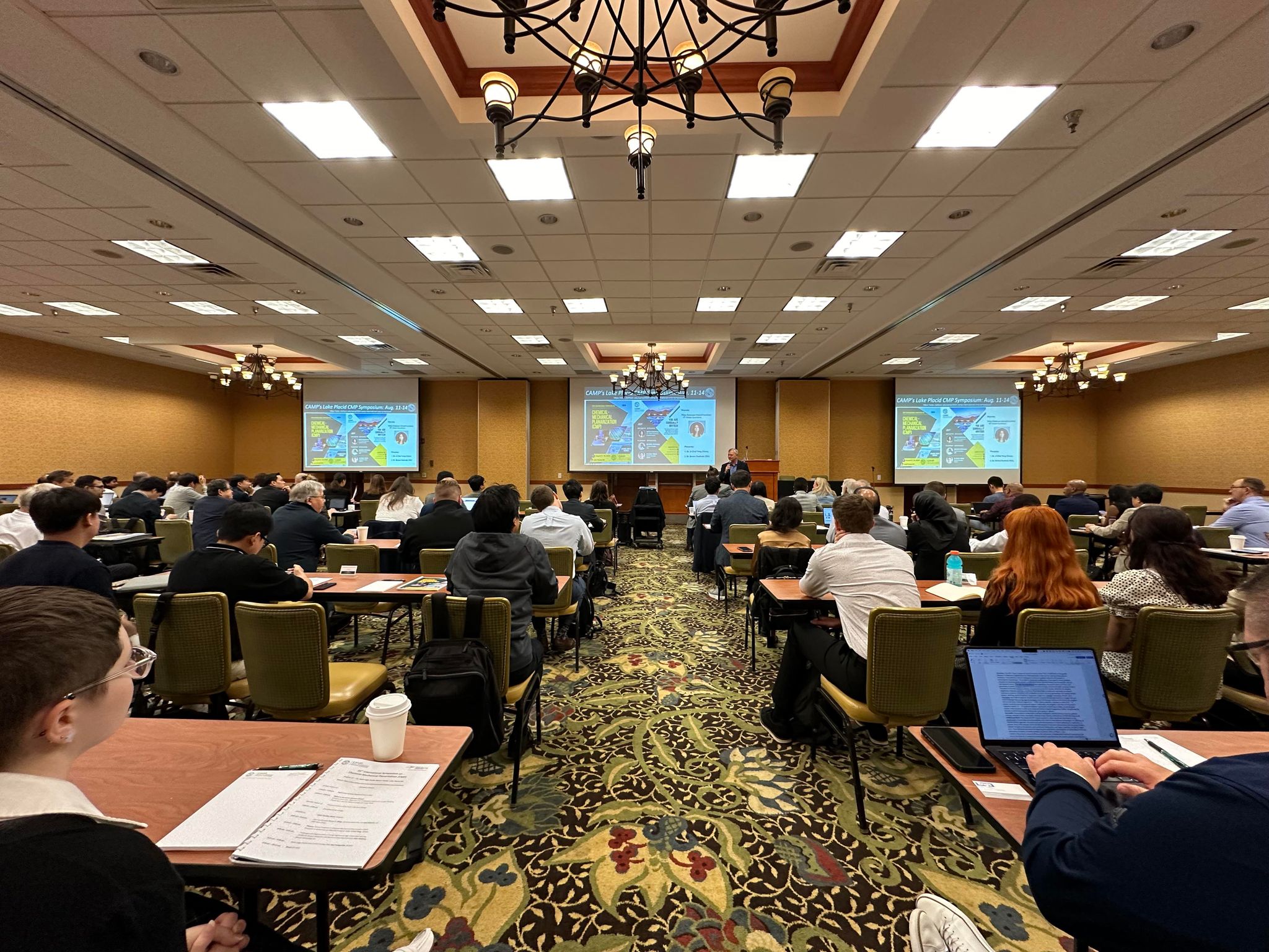 Conference attendees sit facing away, looking toward projector screens in a conference room toward projector screens where a presentation is being given. 