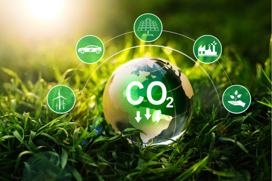 A globe with 'CO2' and downward arrows overlaid, surrounded by icons representing sustainable energy sources such as solar panels, wind turbines, electric vehicles and green manufacturing, set against a background of lush grass in sunlight.