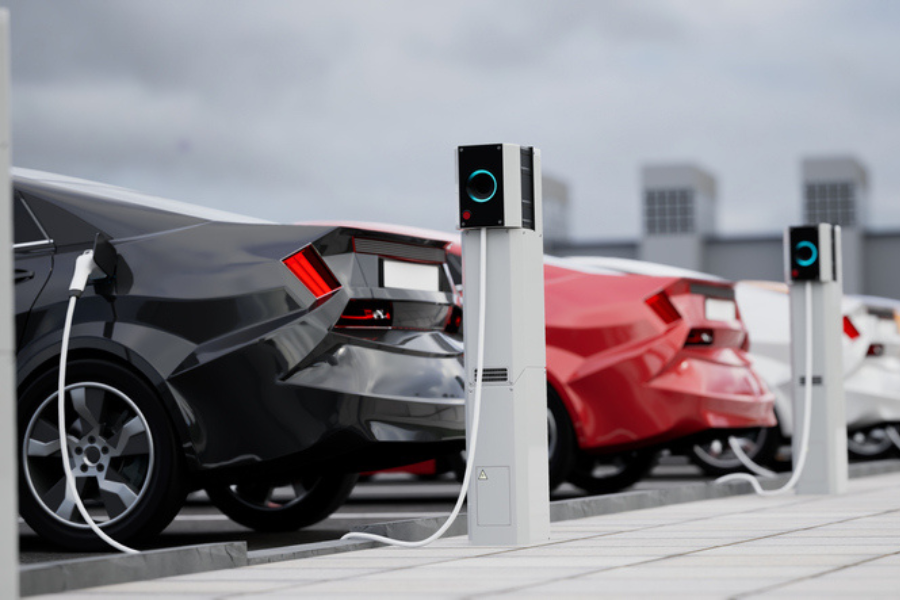 Several electric vehicles, including black, red, and white cars, connected to charging stations in a parking lot under a cloudy sky. The image emphasizes the growth of electric vehicle infrastructure.