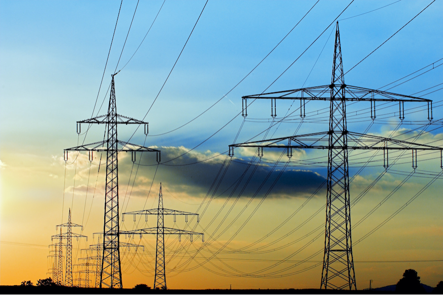 Rows of high-voltage power lines stretching across the landscape during sunset, with a clear sky transitioning from light to darker shades. The image emphasizes energy infrastructure and transmission against a natural backdrop.