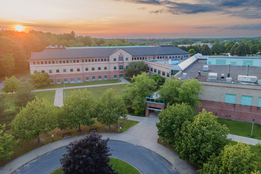 Aerial view of the Center for Advanced Material Processing (CAMP) at Clarkson University during sunset, with the building surrounded by trees and a circular driveway in the foreground. The warm colors of the sunset highlight the campus landscape, emphasizing the scenic environment.