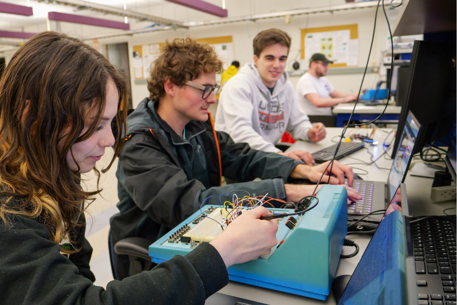 Clarkson University students working collaboratively in an electrical engineering lab, using equipment and laptops to build and test circuits. The environment showcases hands-on learning and teamwork in a technical academic setting.