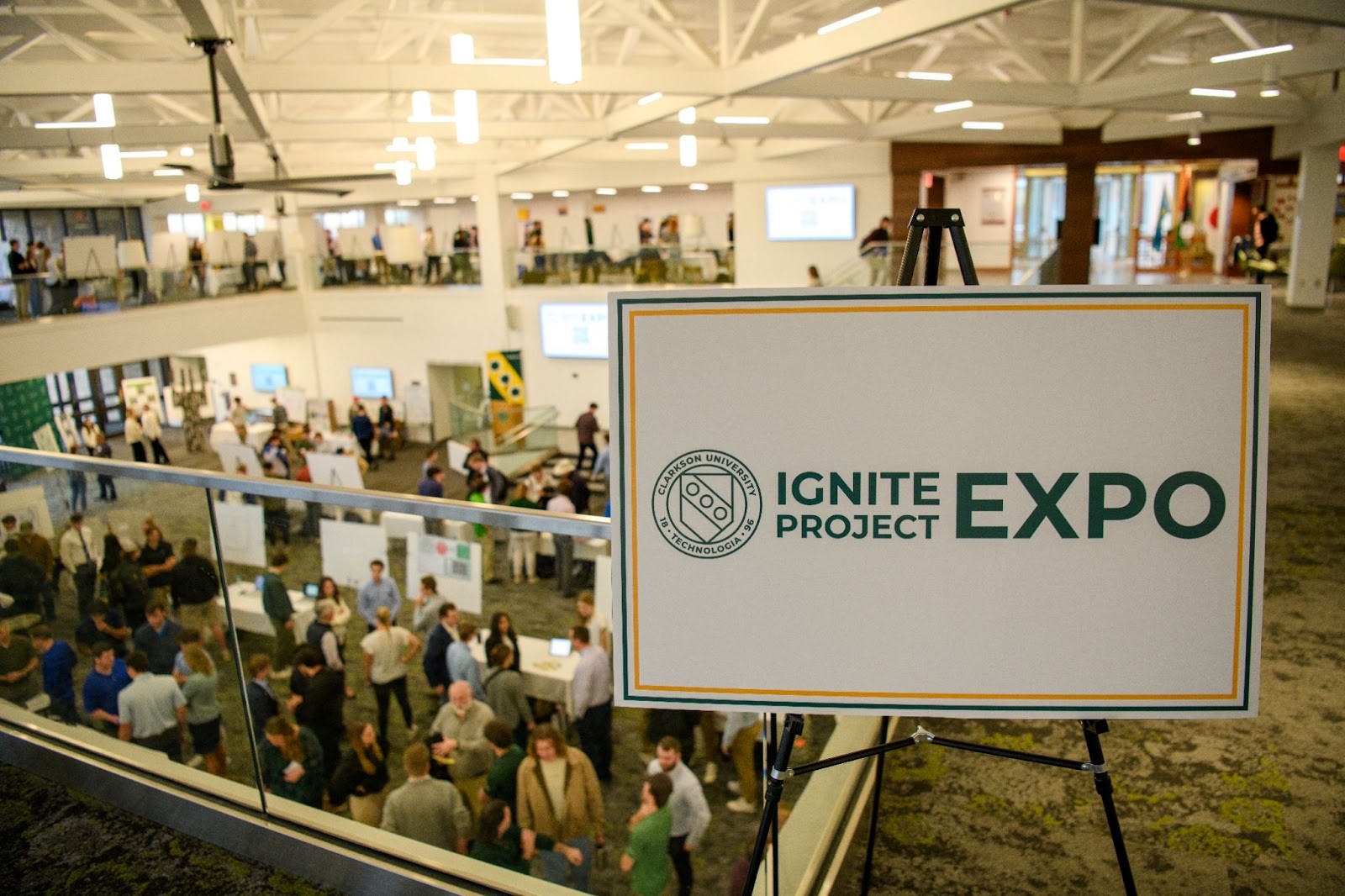 White sign on an easel with the Clarkson University logo and "Ignite Project Expo" written on it faces the camera while an atrium full of students with posters at tables fills the background. 