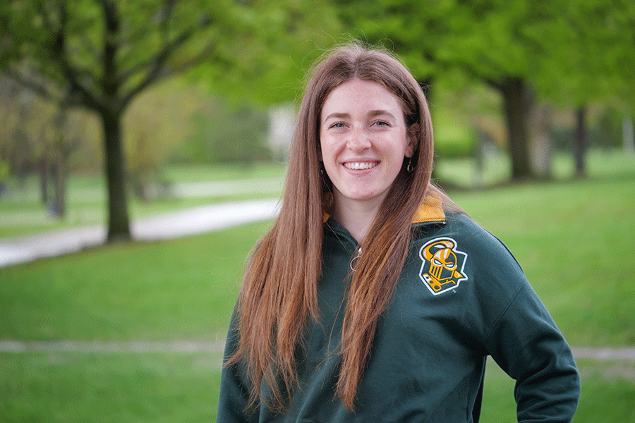 Clarkson Ignite graduate fellow Kelsey Hamilton poses outside on a sunny day in front of trees while wearing a Clarkson athletics logo sweatshirt