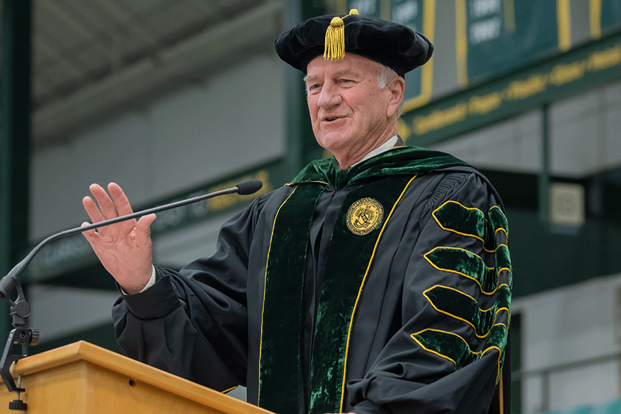 Clarkson President Emeritus Tony Collins wears regalia and speaks at a lectern during Clarkson's annual Commencement Ceremony in Cheel Arena