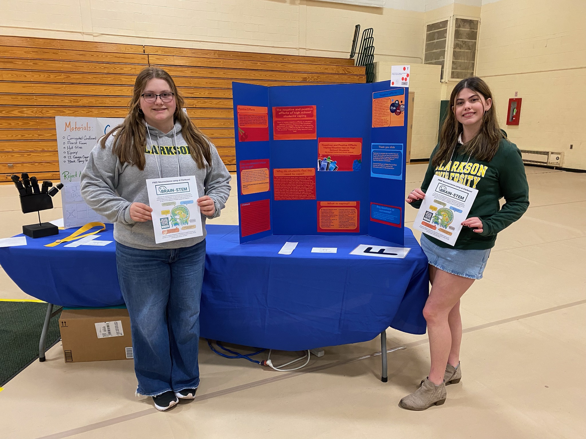 Two girls stand on either side of a science fair poster, holding certificates