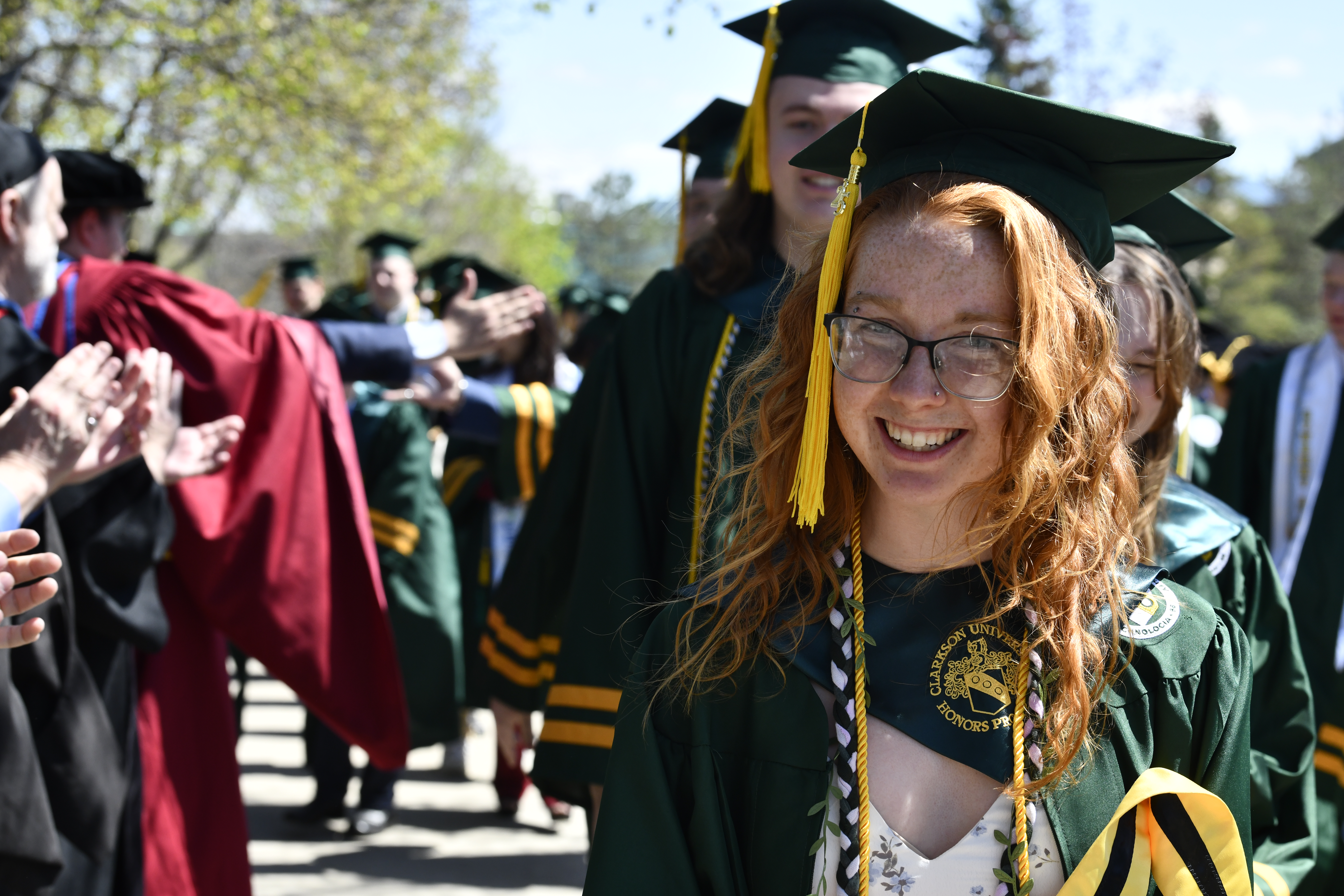 A young woman in regalia smiles at the camera as she walks outdoors ahead of commencement