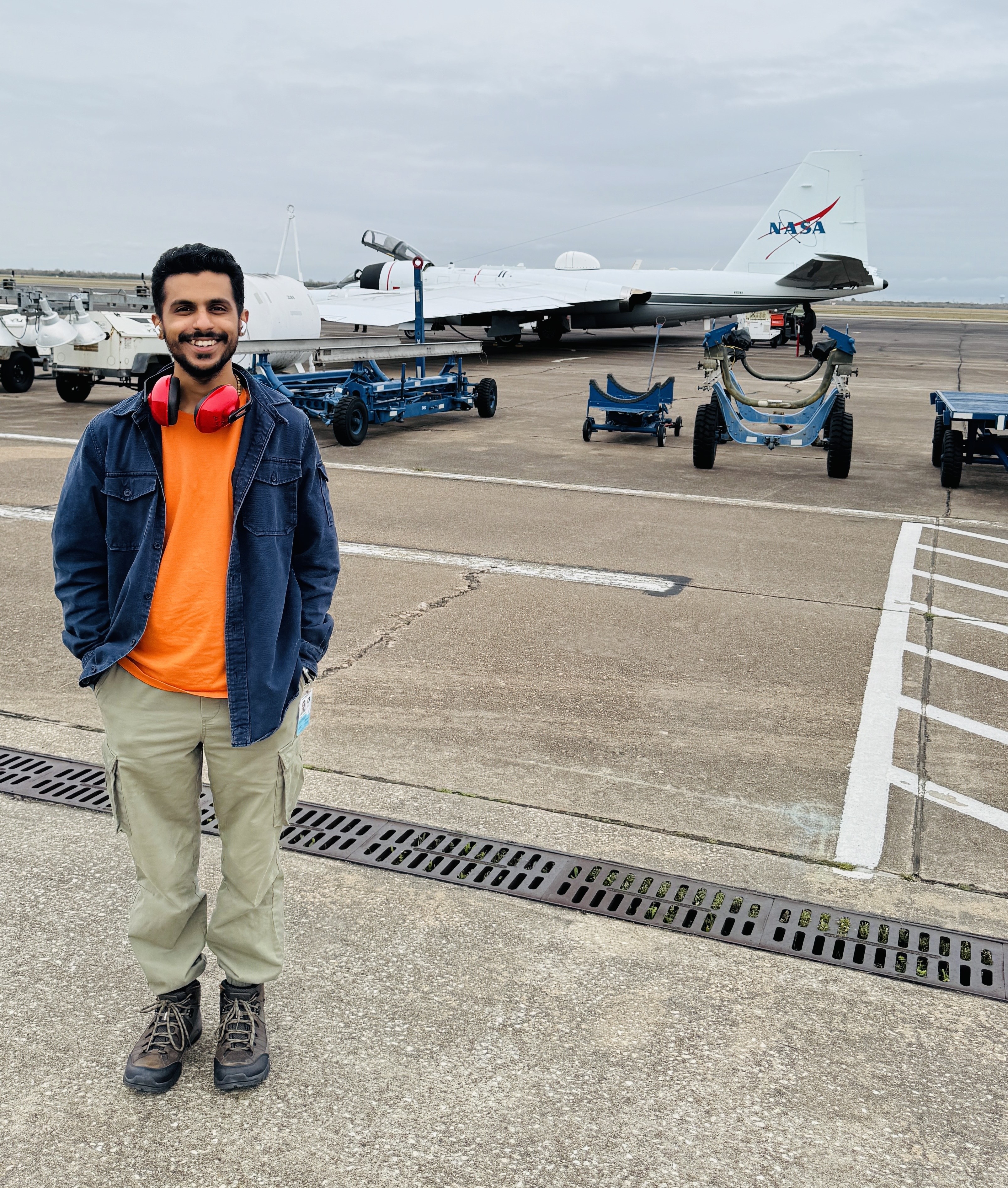 A young man smiles for a photo on a tarmac with a plane and other runway equipment in the background.