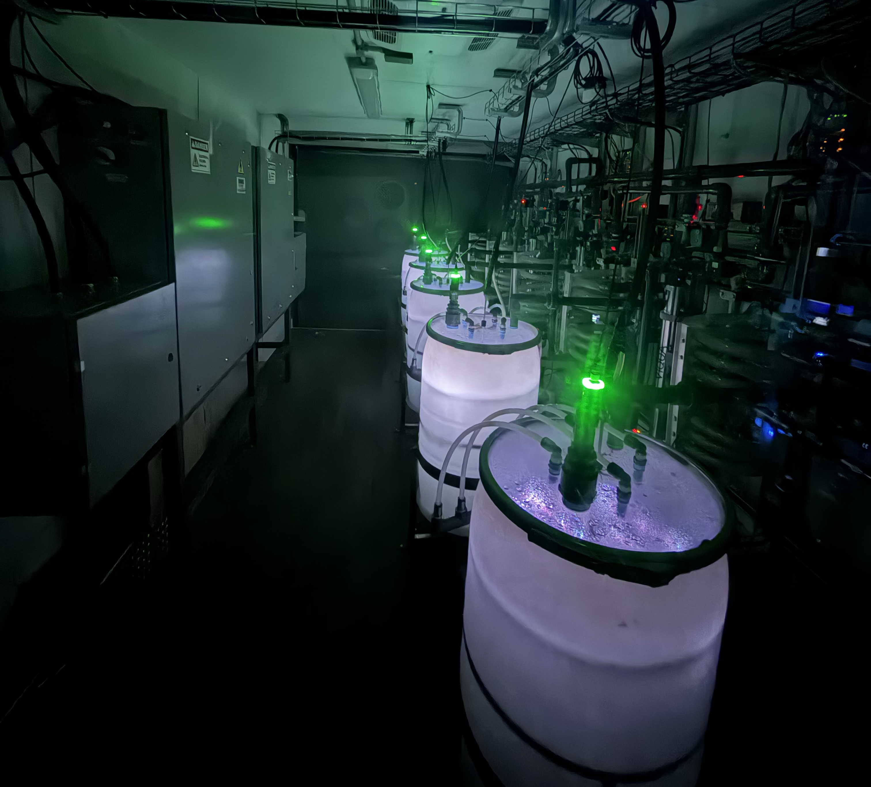 55 gallon plastic drums with glowing green lights attached to the lid sit in a row in a dark room