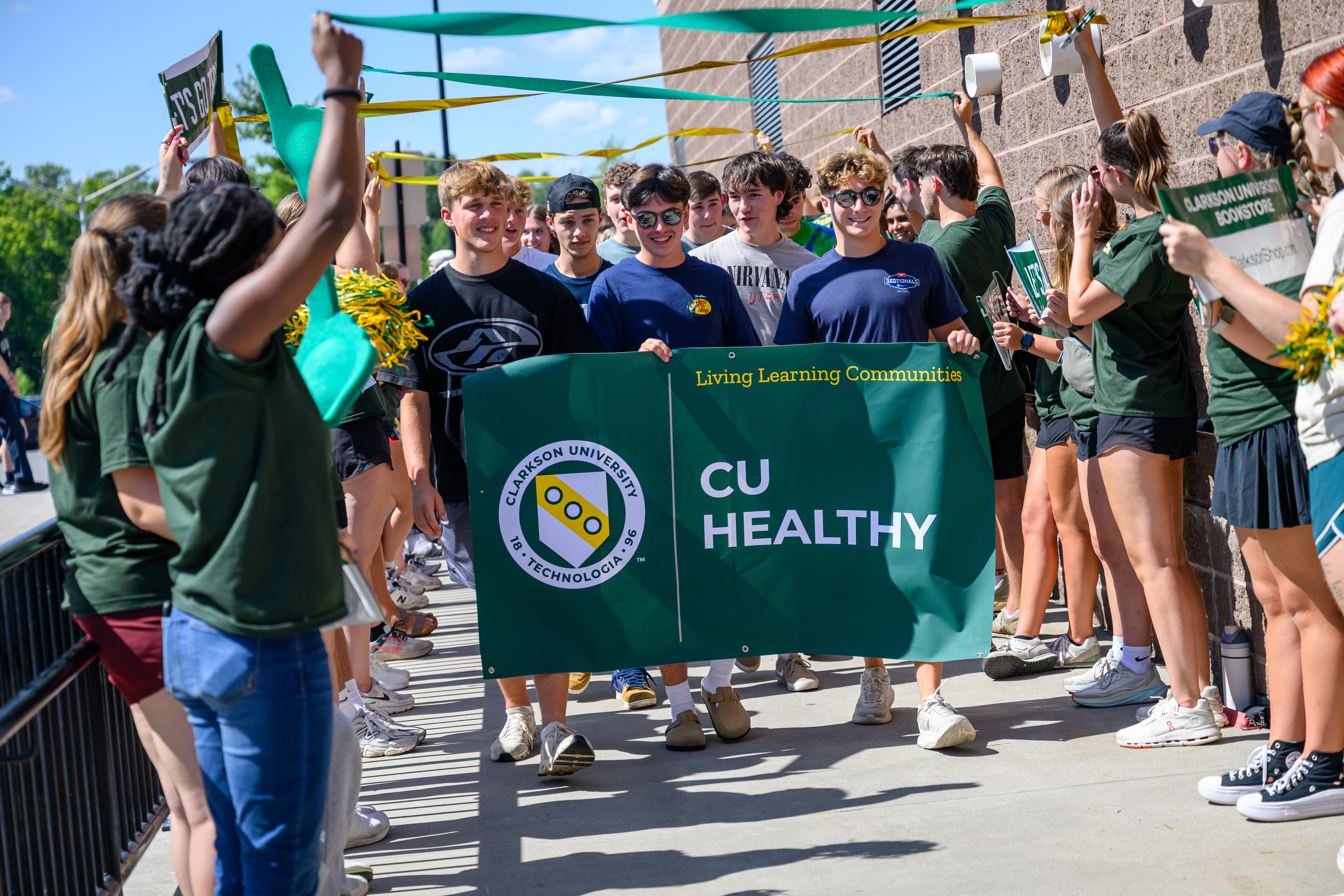 Clarkson University freshman students walk through a cheering crowd during freshman convocation, carrying a banner that reads “CU Healthy” with the Clarkson University logo while other students line the walkway holding signs and streamers.