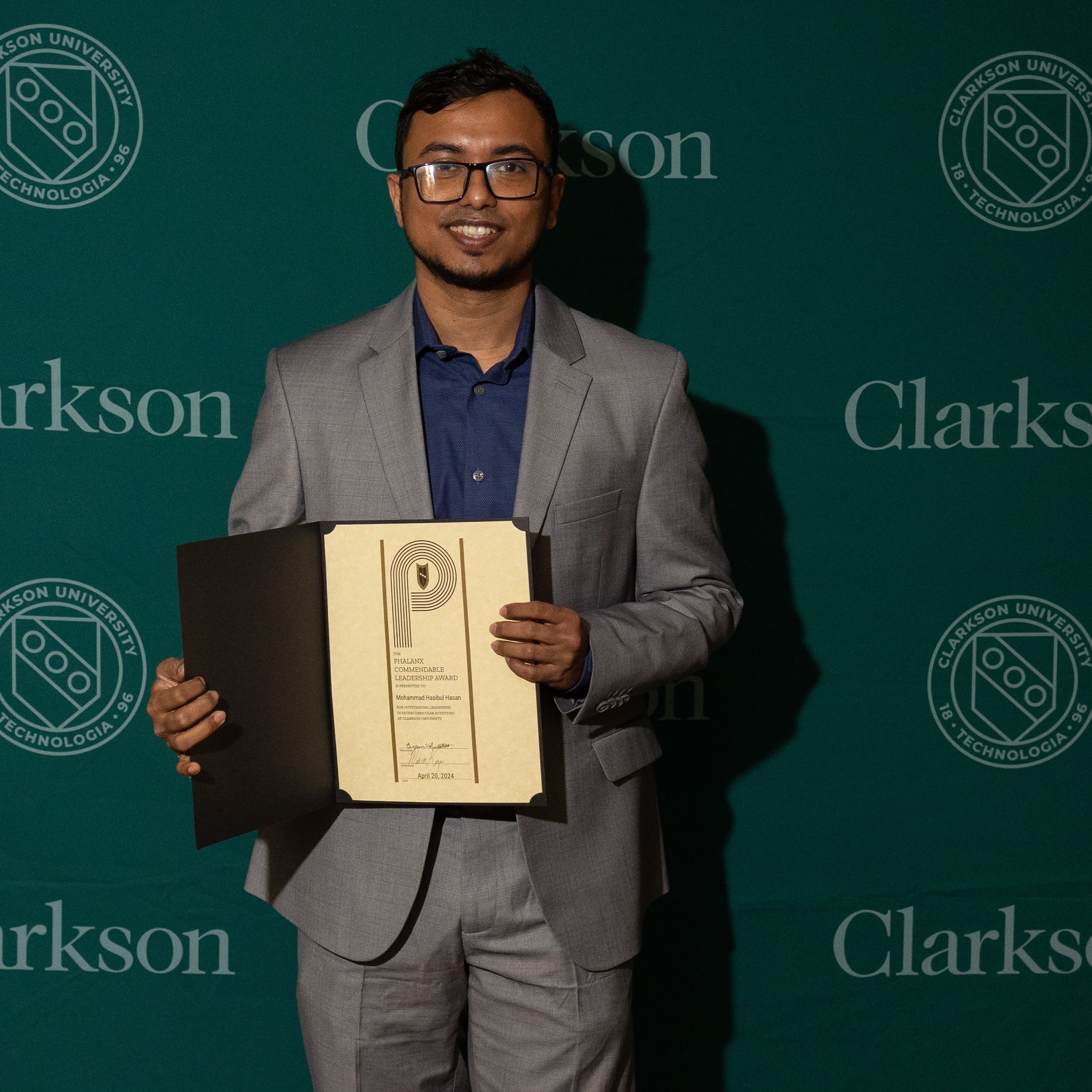 Mohammad Hasibul Hasan smiling while holding an award. He is wearing a grey suit and standing in front of a green Clarkson University backdrop.