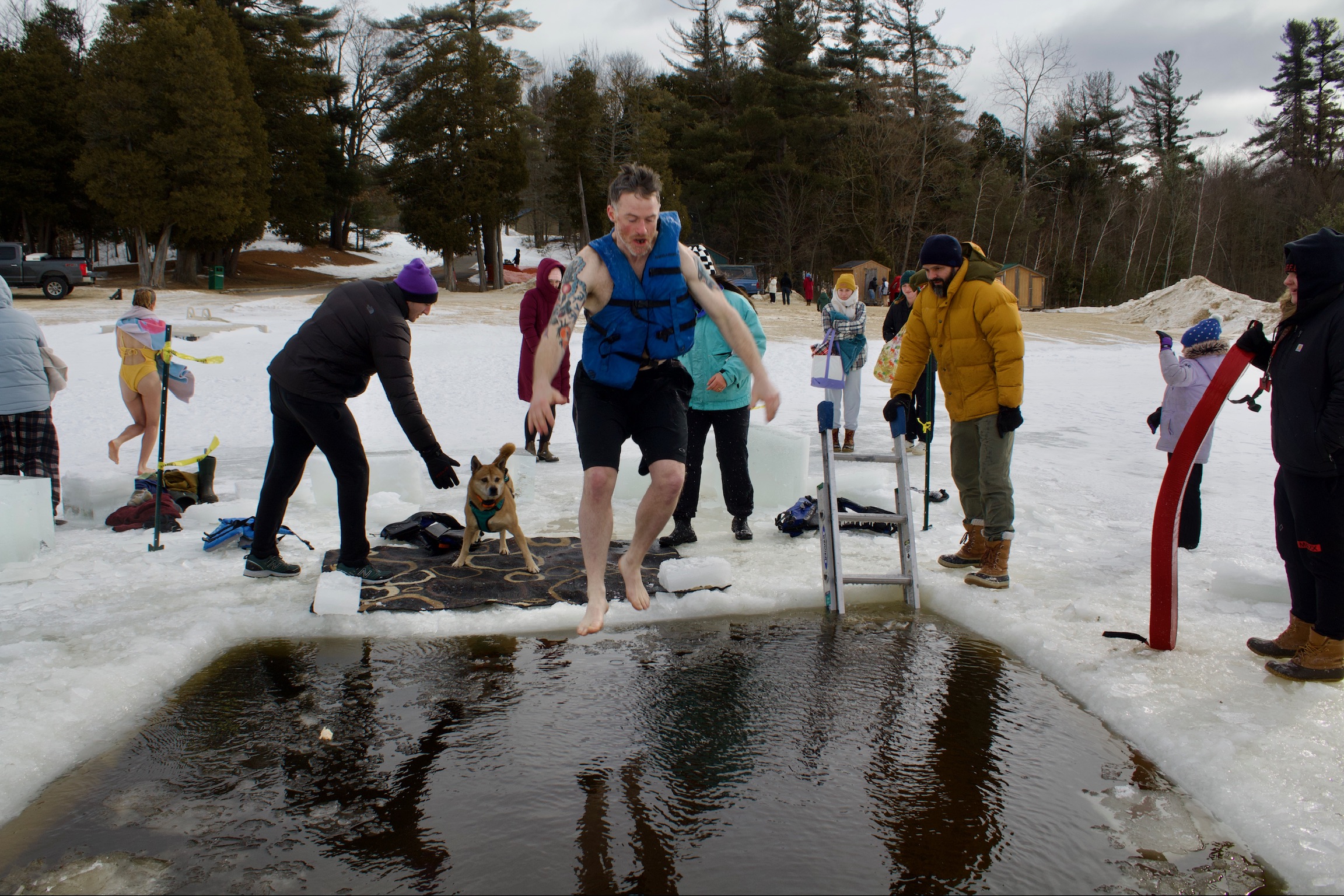 A man jumps into a hole cut in the ice on a body of water in winter, surrounded my volunteers from the event.