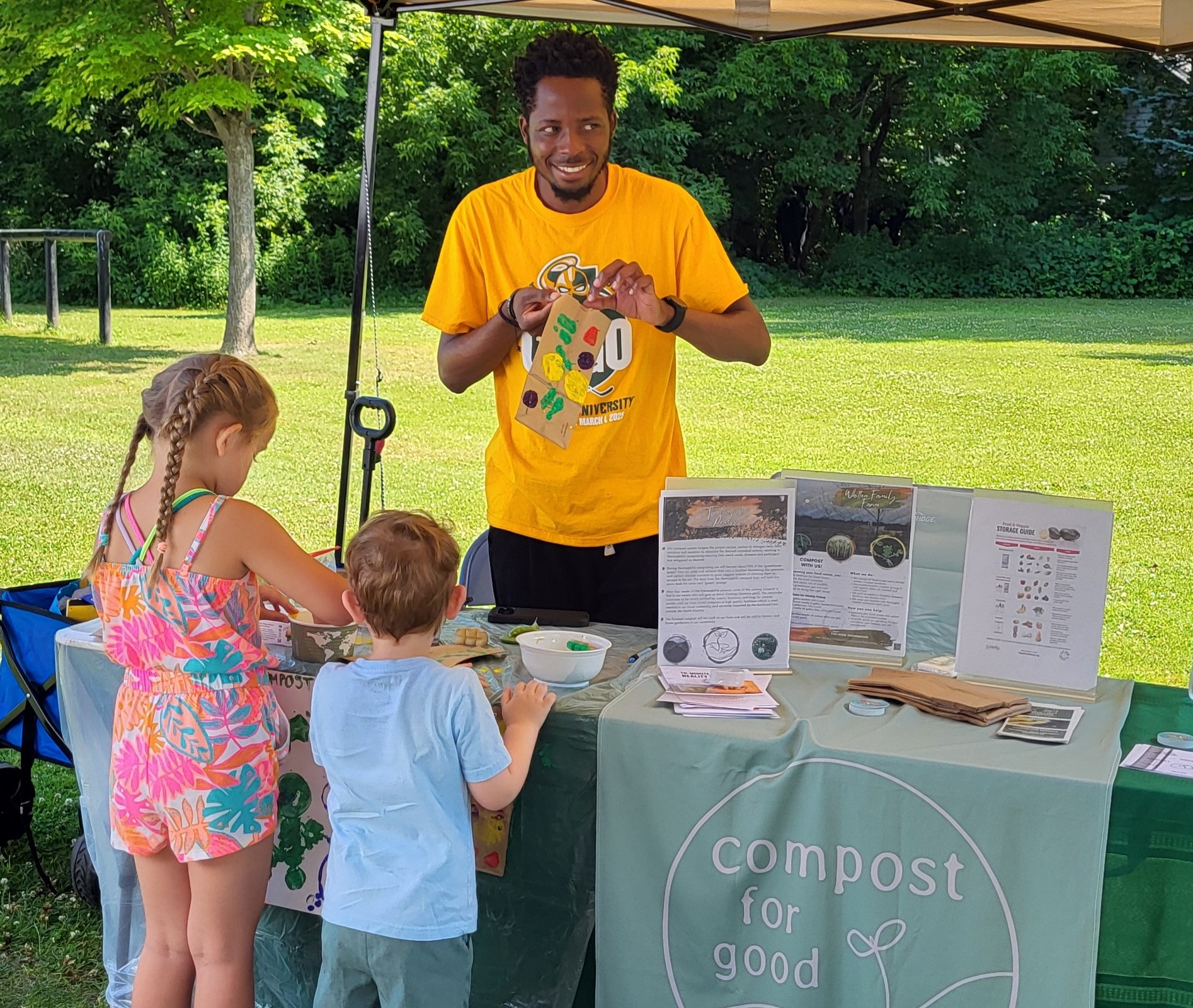 A young man tabling for Compost for Good holds a child's art project, while two children peruse items on the table. 