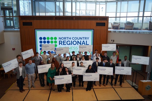 Students stand on a stage with oversized checks representing their awards at the North Country Regional Business Development Competition