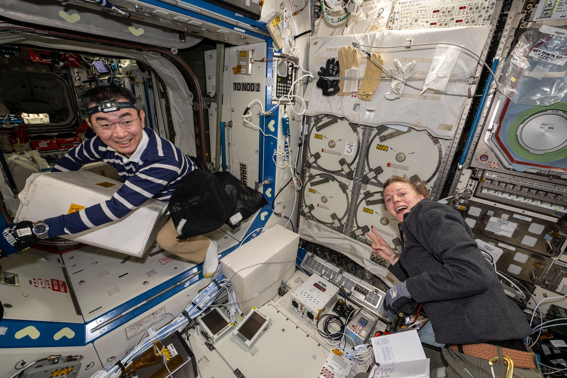 Two astronauts inside a spacecraft module surrounded by equipment, cables, storage panels, and control systems in a microgravity environment. One astronaut on the left smiles while handling a large white container, while the astronaut on the right smiles toward the camera and makes a peace sign gesture.