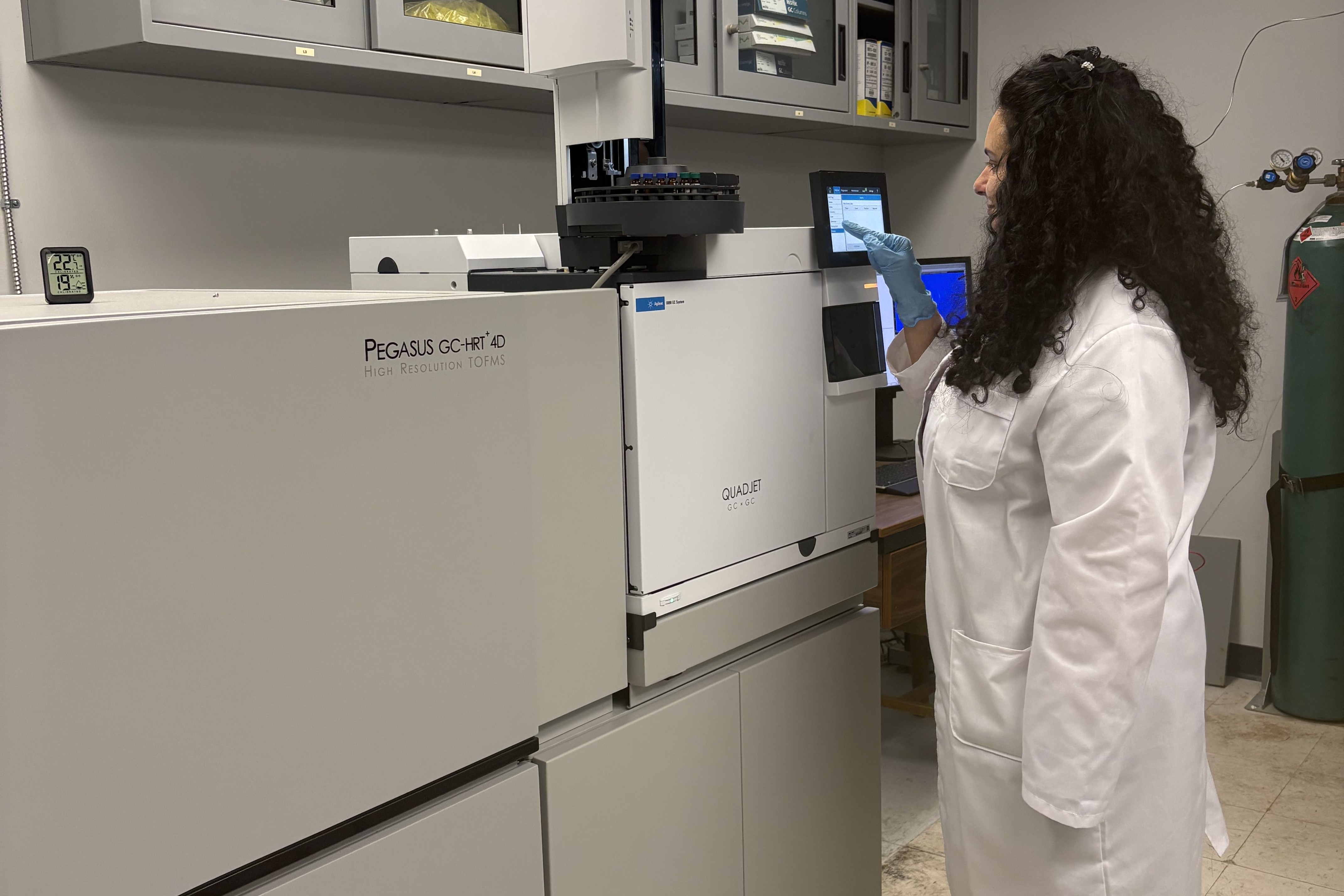 A female Clarkson University researcher with curly hair, wearing a white lab coat and blue gloves, operates the touch screen interface of an Agilent gas chromatography system. A larger analytical instrument and laboratory storage cabinets are visible in the background.