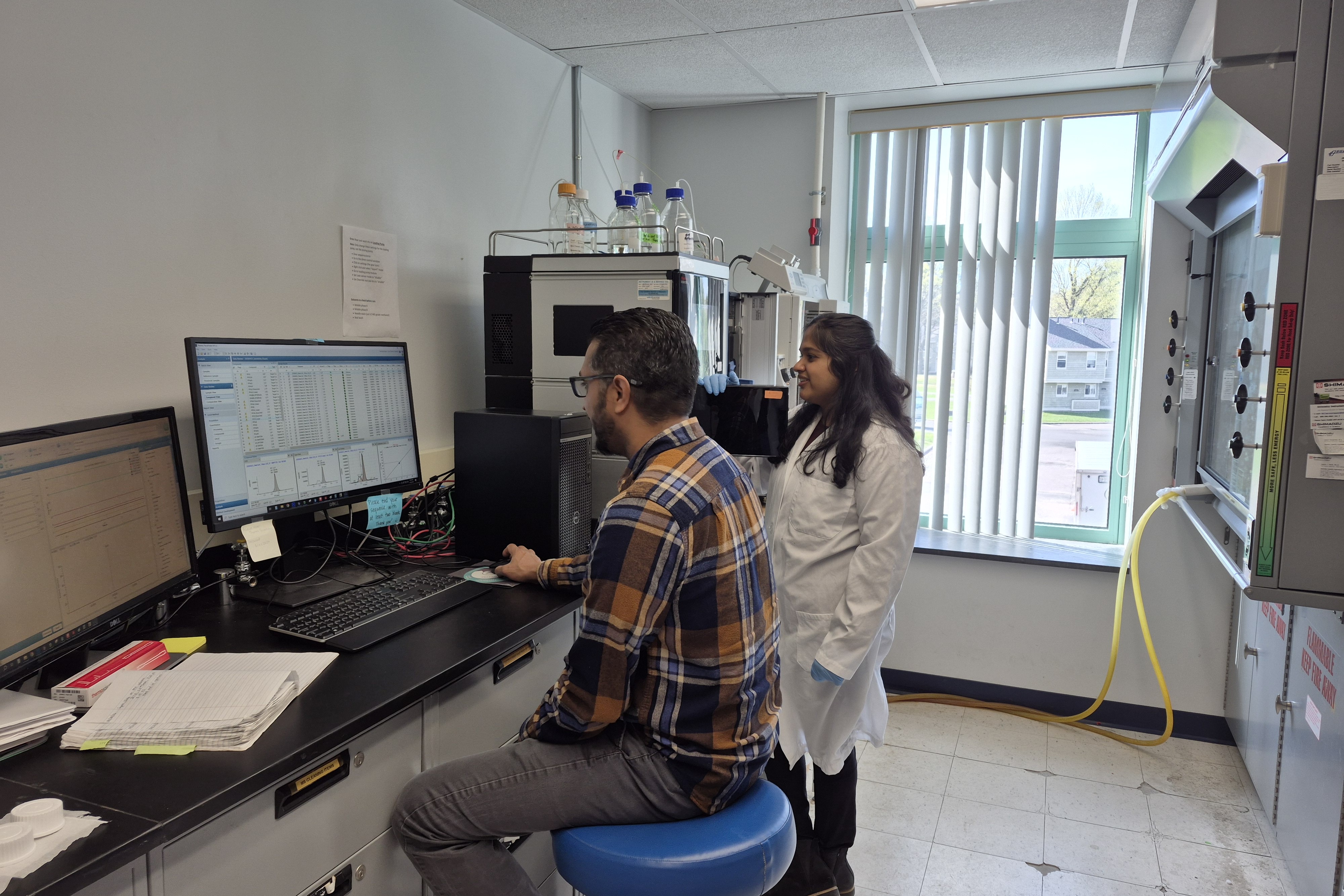 Two Clarkson University researchers analyzing data in a laboratory setting. A male researcher sits at a computer desk looking at complex charts on a monitor, while a female researcher in a white lab coat stands nearby, looking on. High-performance liquid chromatography (HPLC) instrumentation and a laboratory fume hood are visible in the background.