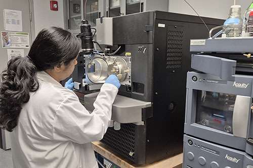 A researcher wearing a white lab coat and blue gloves adjusts the open ion source chamber of a large mass spectrometer in a university laboratory.