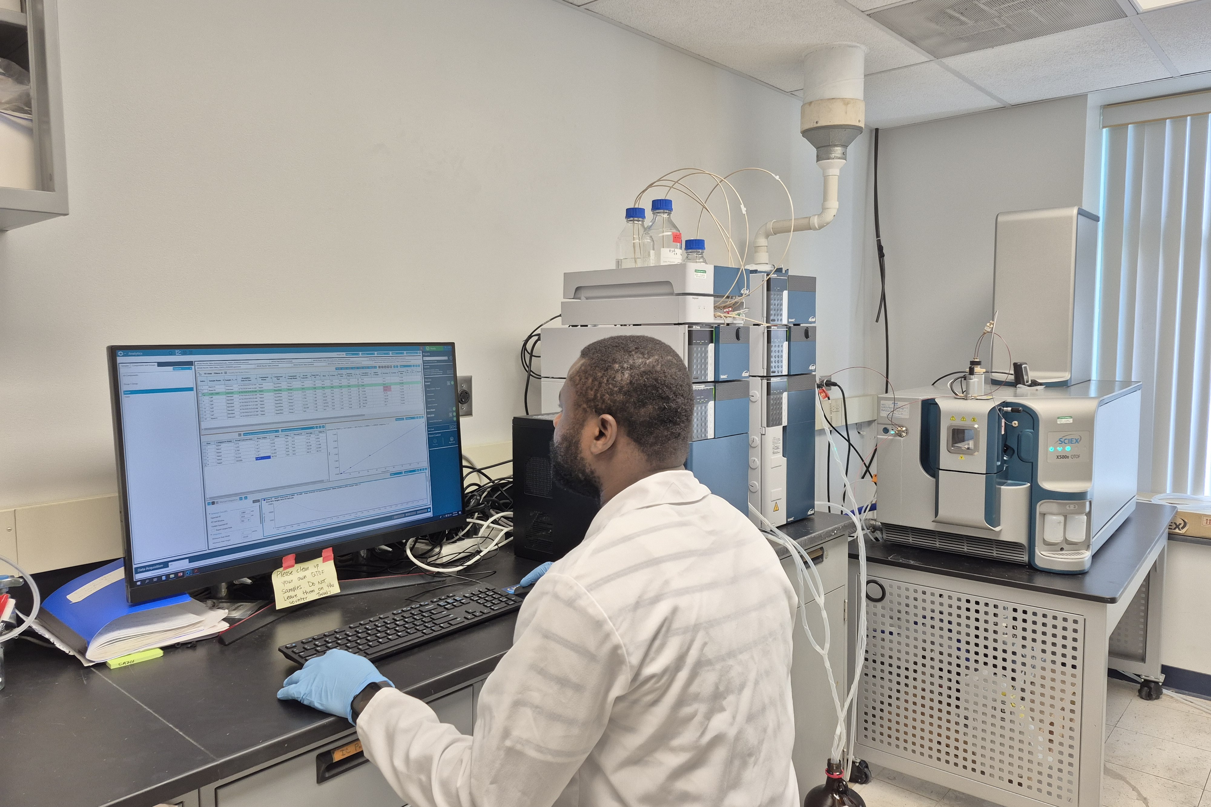 A Clarkson University researcher in a white lab coat and blue gloves sits at a computer terminal analyzing analytical software. In the background, a high-performance liquid chromatography (HPLC) system is coupled with a mass spectrometer on a laboratory workbench.