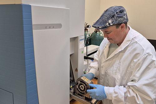A researcher wearing a flat cap, white lab coat, and blue gloves works on a component of a large analytical mass spectrometer in a university lab.
