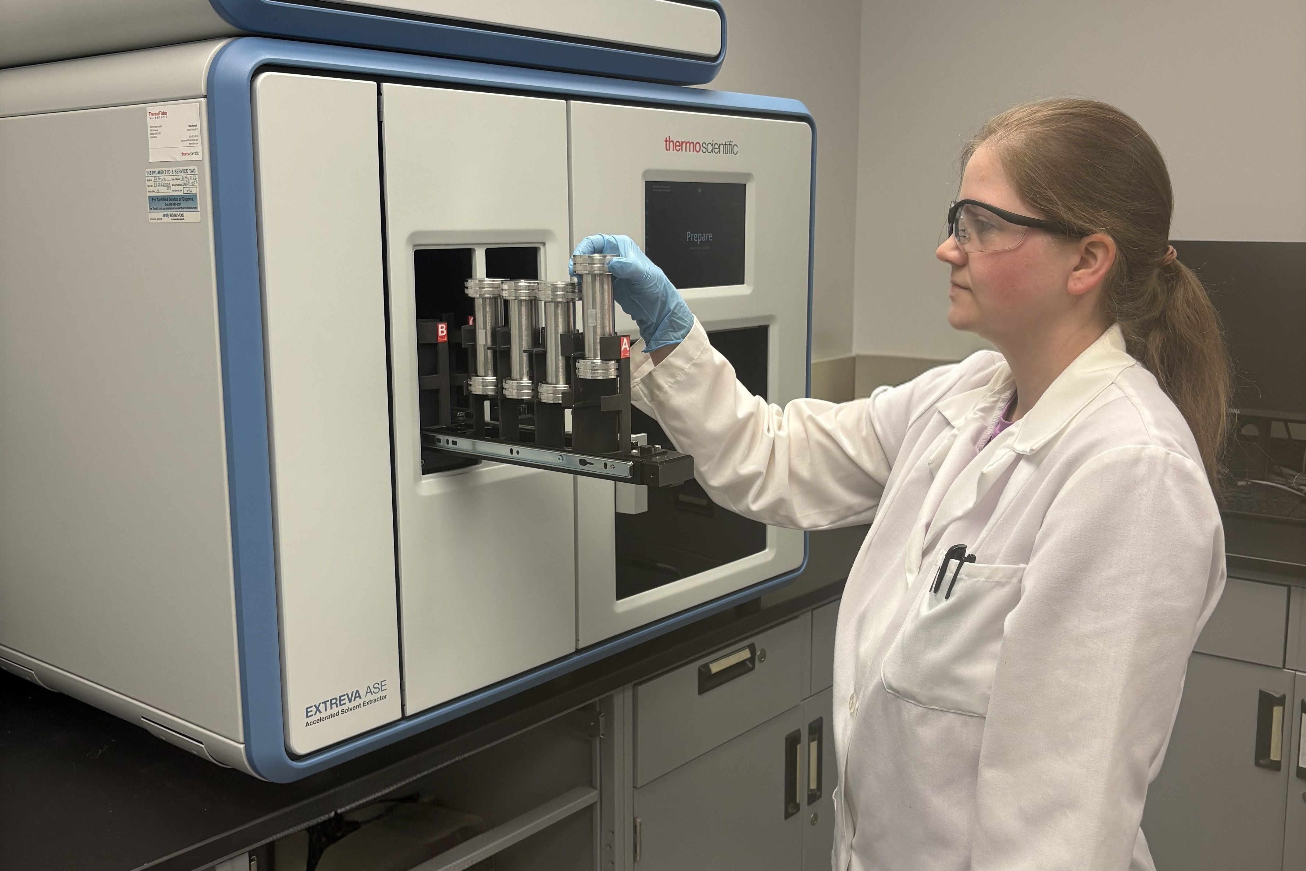 A female Clarkson University researcher wearing safety glasses, a white lab coat, and blue gloves adjusts metal sample cells on a tray protruding from a Thermo Scientific Dionex ASE (Accelerated Solvent Extractor) system on a laboratory countertop.