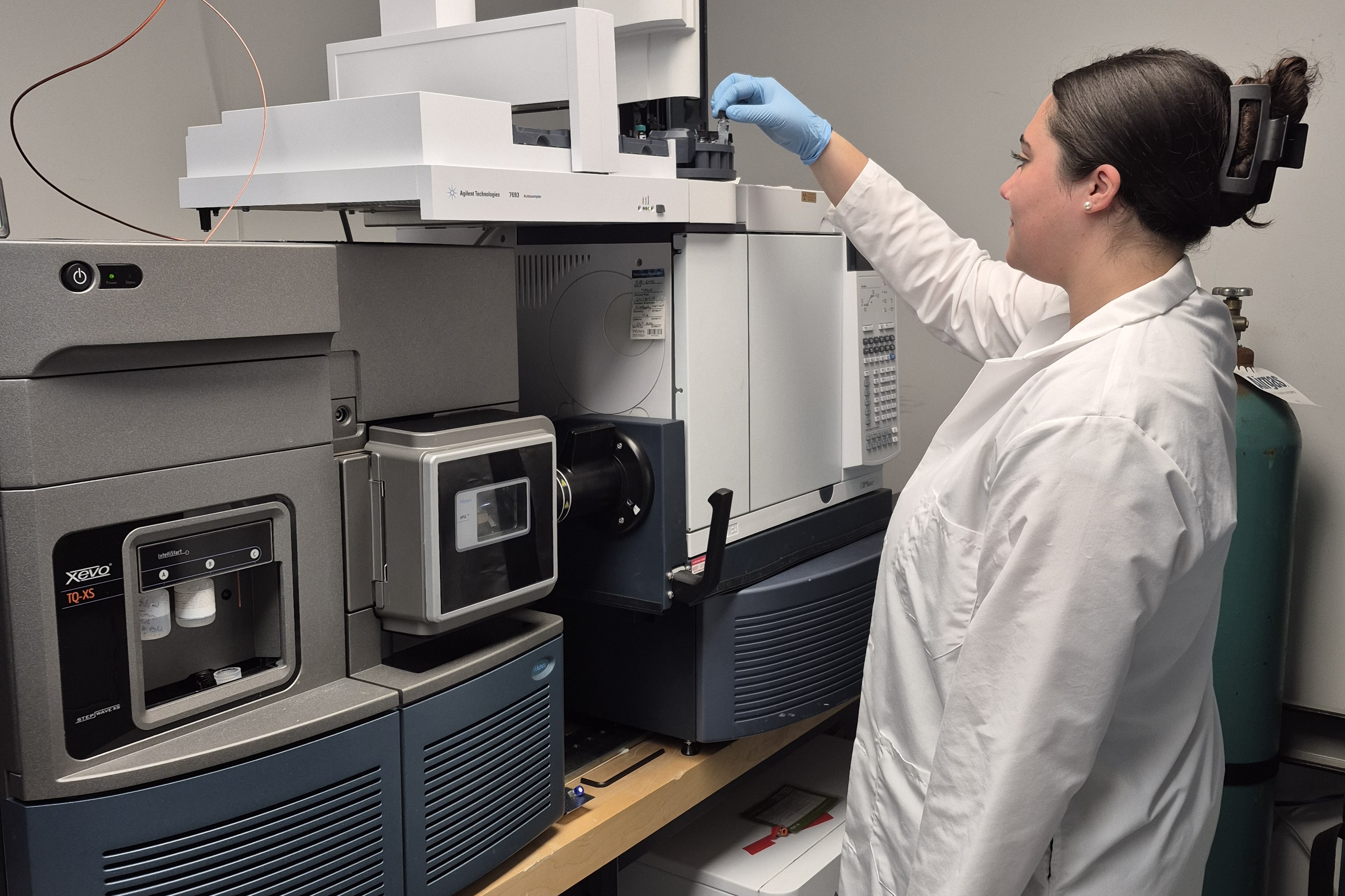 A female Clarkson University researcher wearing a white lab coat and blue gloves loads a sample into an automated autosampler. In the foreground, an advanced mass spectrometry and gas/liquid chromatography system sits on a laboratory workbench.