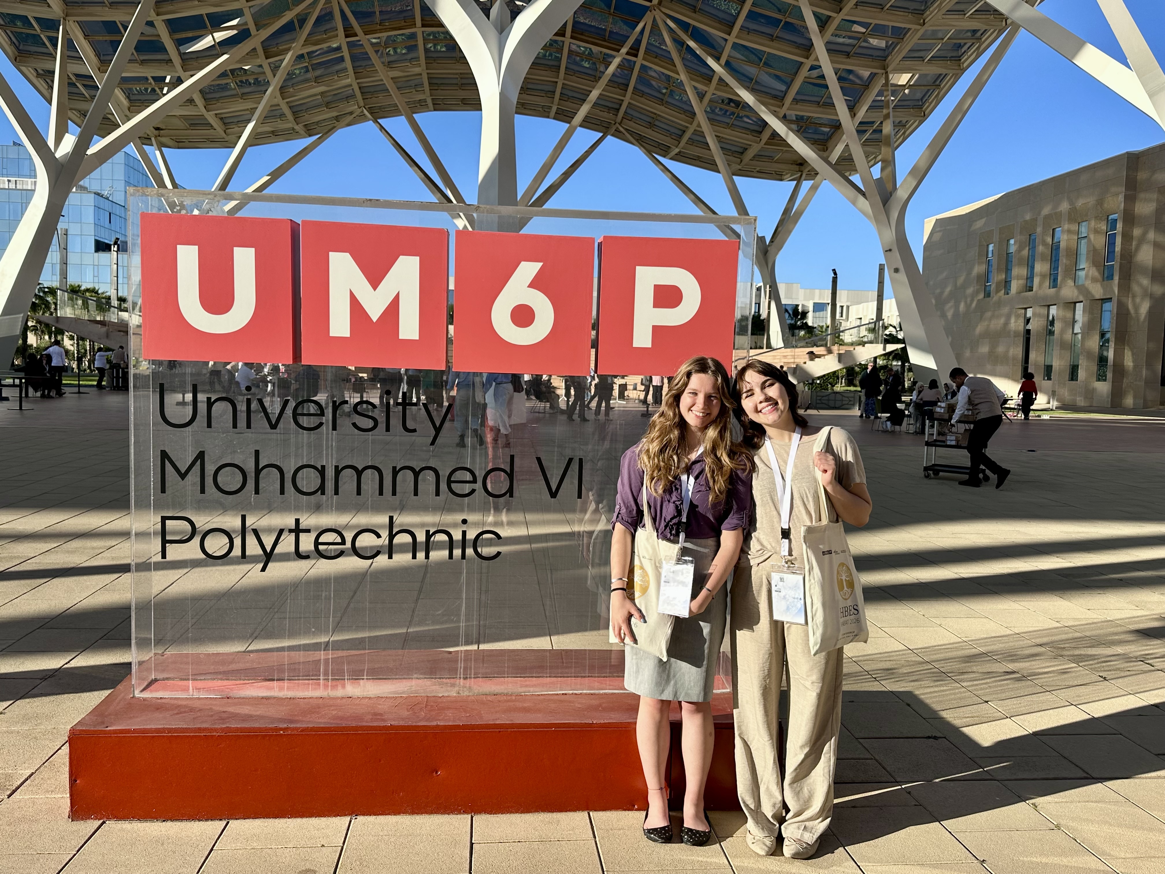 Two students stand smiling in front of a large sign for University Mohammed VI Polytechnic (UM6P) on the university's campus. Both are wearing conference badges and carrying tote bags, suggesting they are attending an academic event. The modern campus features a striking steel canopy overhead, contemporary buildings, and people walking through the plaza in the background under a clear blue sky.