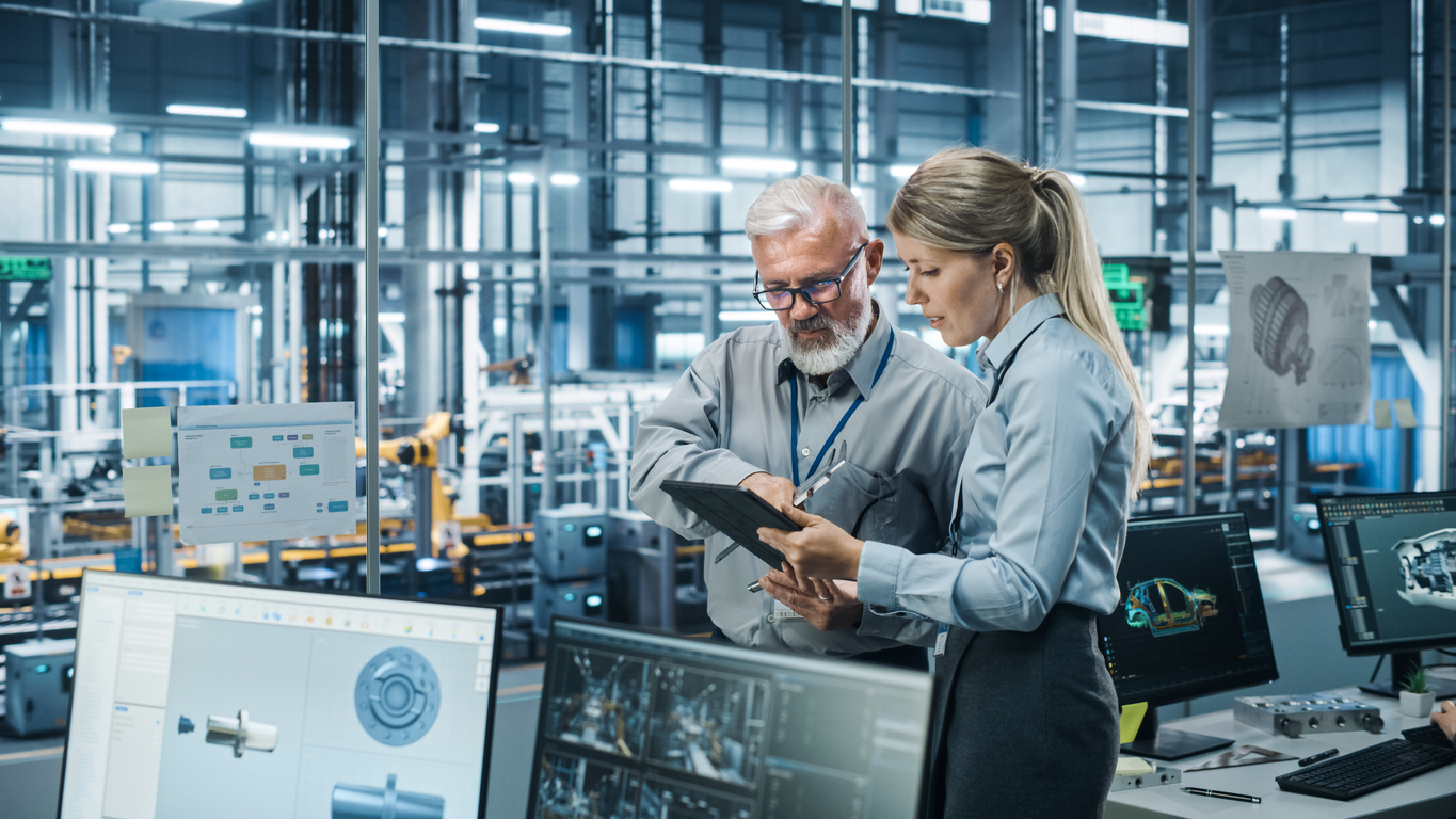 Two engineers stand in a modern manufacturing facility reviewing a tablet together, with computer monitors displaying 3D design models and automated production equipment visible in the background.