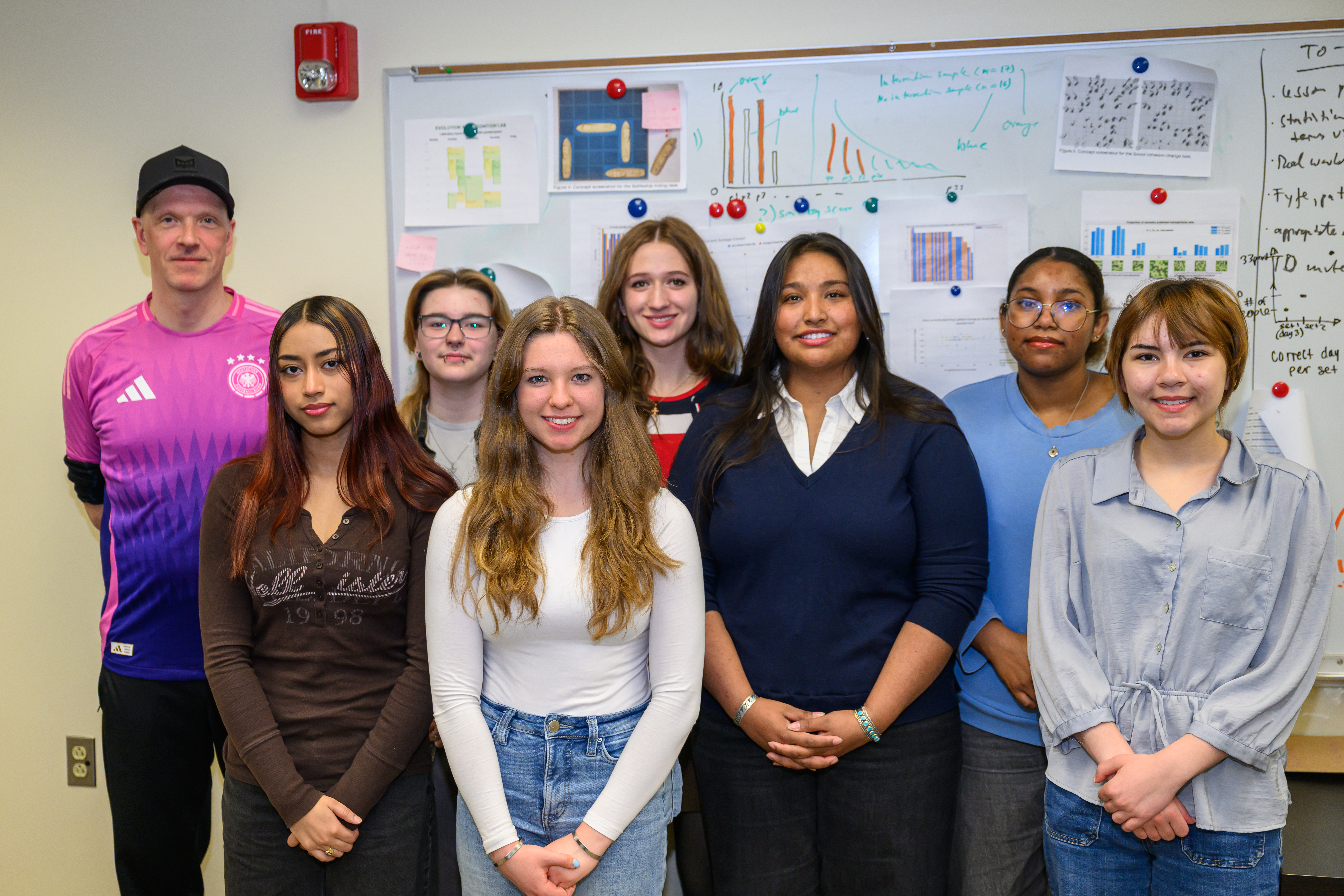 A group of eight Clarkson University students posing together in a classroom in front of a whiteboard covered with project charts and graphs.
