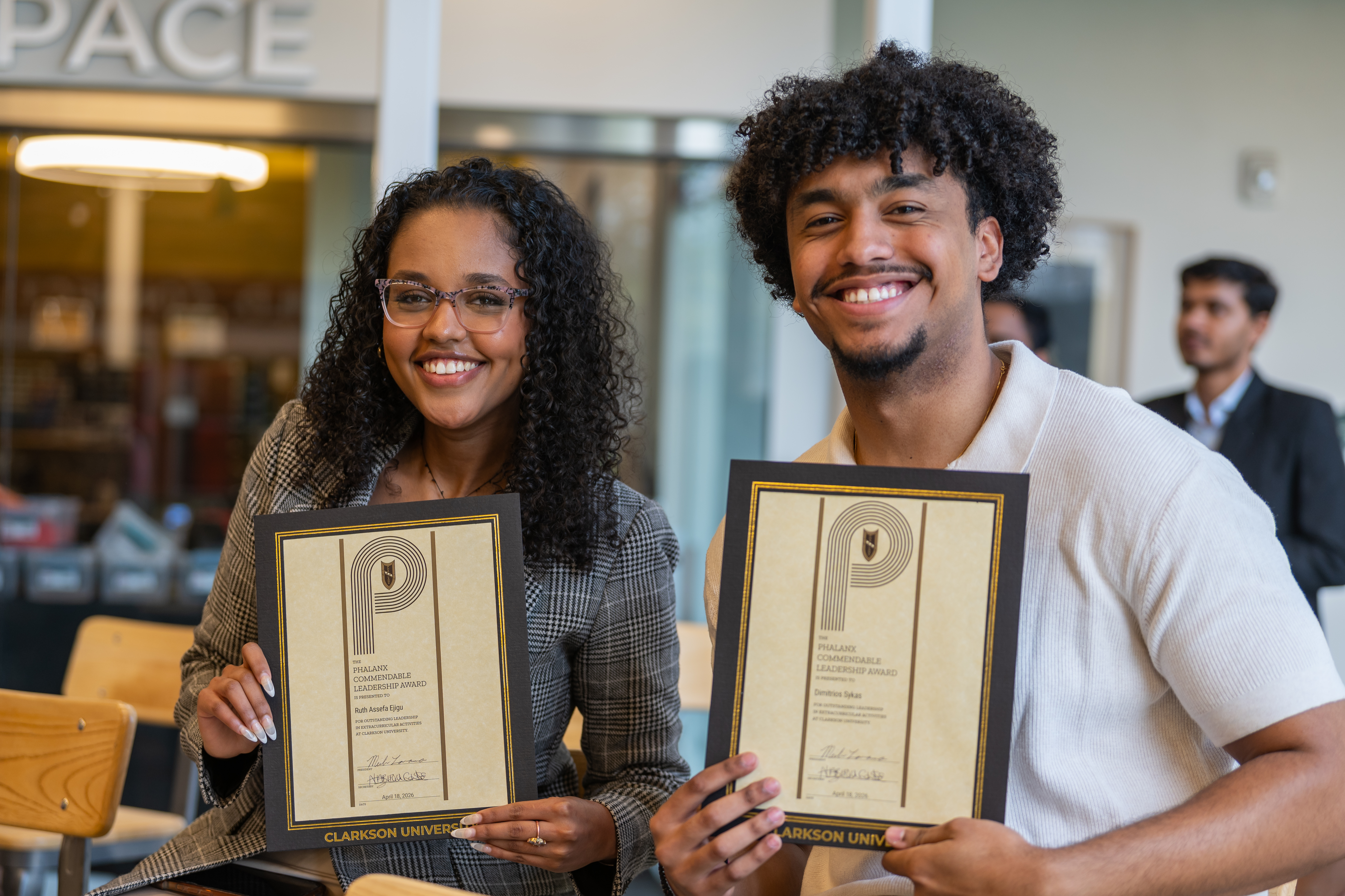 Two smiling Clarkson University students hold up framed Phalanx honor society certificates.