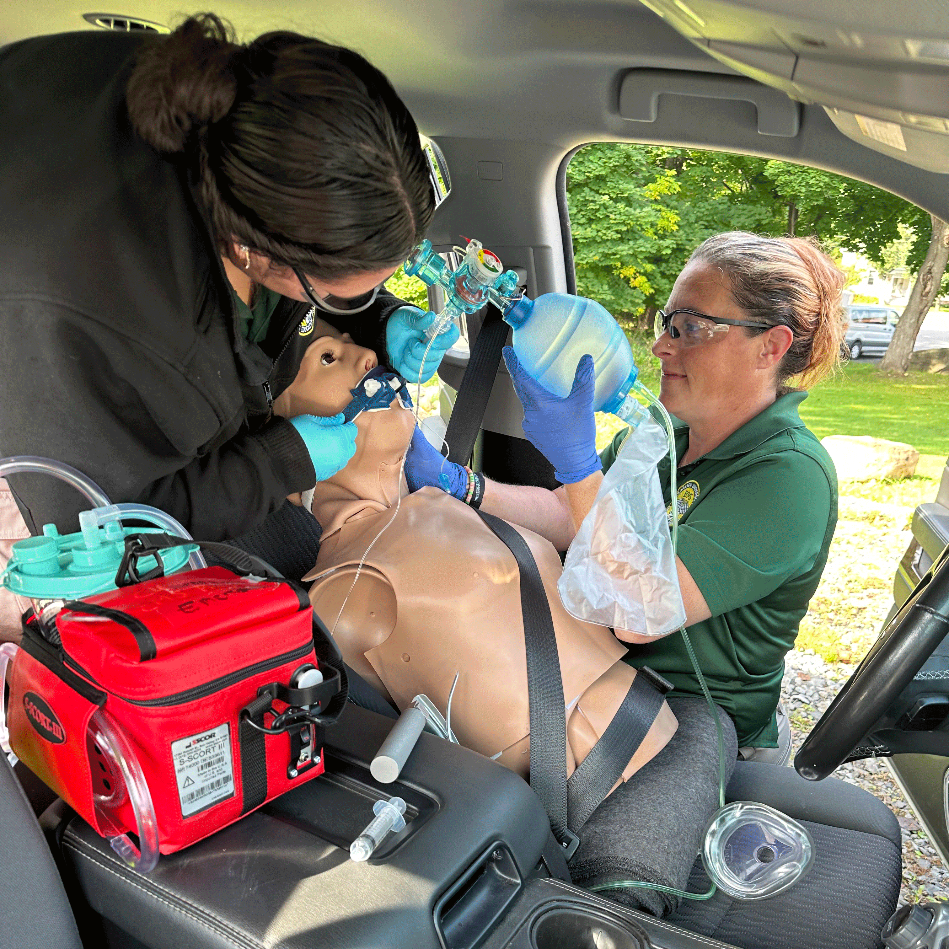 Two students in green Clarkson shirts practice hands-on medical training inside a vehicle. One student secures an oxygen mask while the other operates a bag-valve mask on a mannequin in the driver's seat, surrounded by medical supplies.